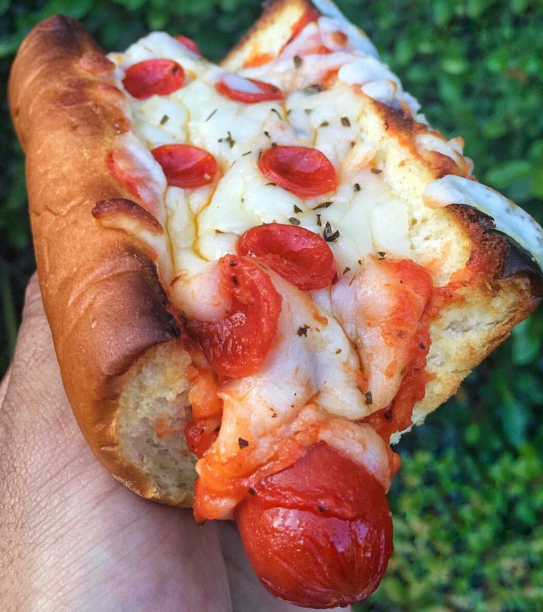 A hand holding a pizza-style hot dog topped with melted cheese, mini pepperoni slices, and herbs. The hot dog bun is toasted and the melted cheese is lightly browned. Greenery is visible in the blurred background.