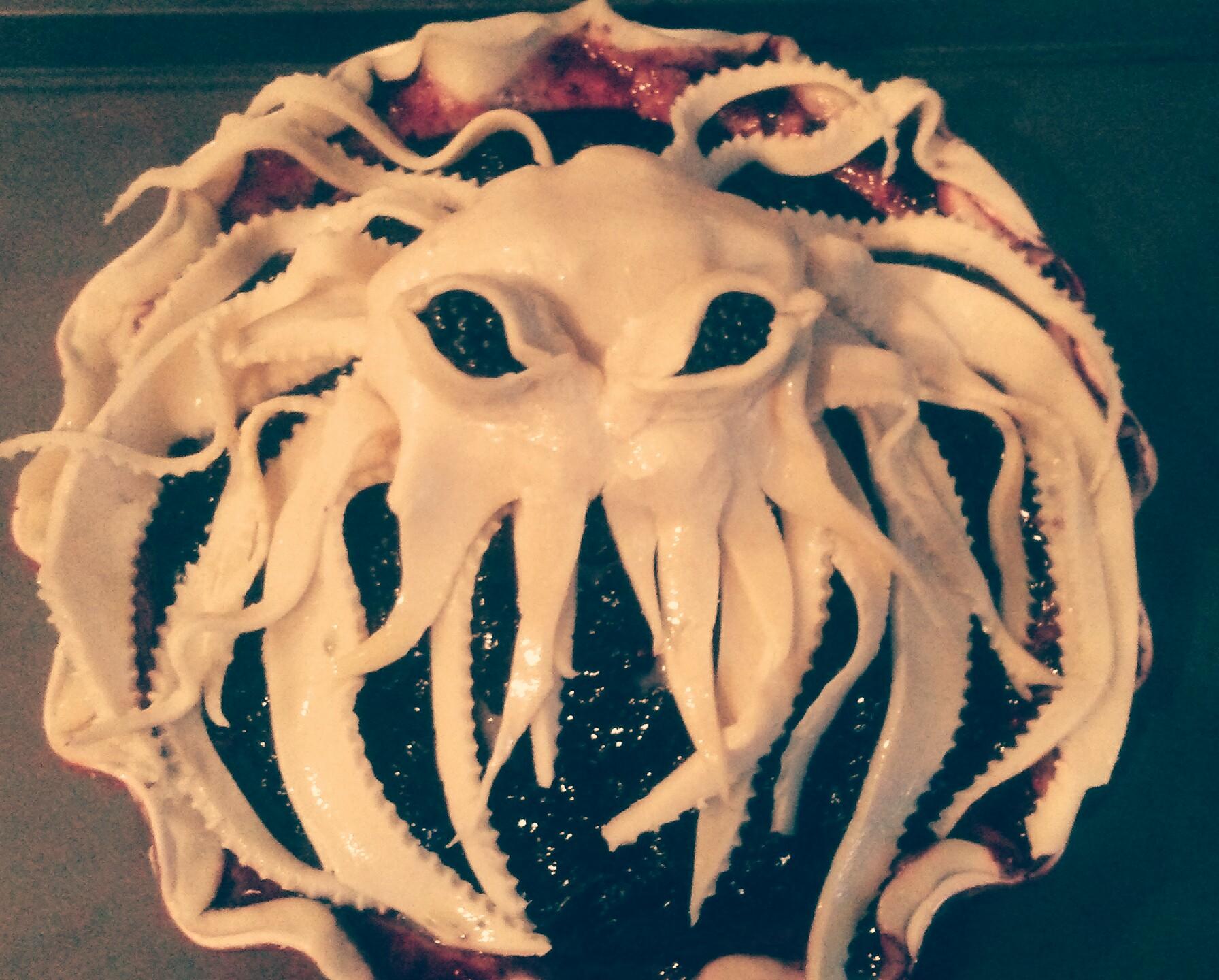 A pie with a top crust shaped like an octopus or sea monster, featuring tentacles and large eye holes, atop a dark fruit filling.