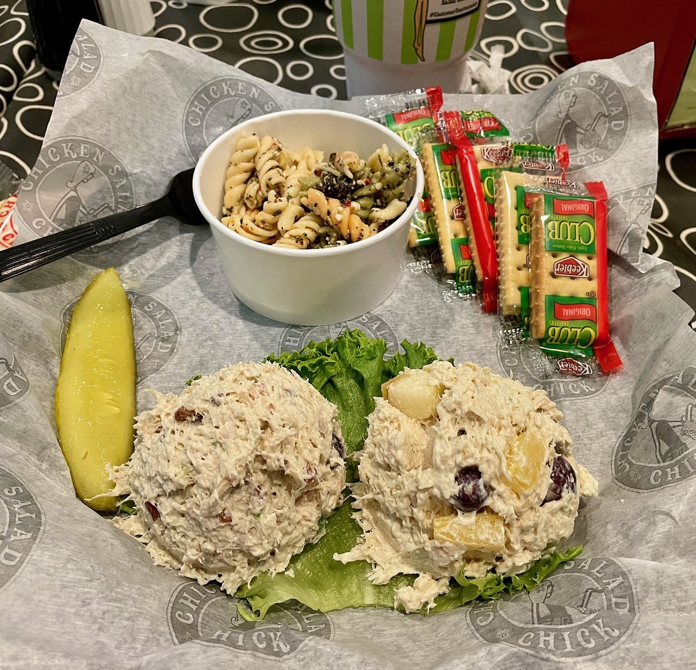 Two scoops of chicken salad on fresh lettuce, a pickle spear, pasta salad in a cup, and packets of Club crackers are served on a tray with Chicken Salad Chick-branded paper—a delicious offering from one of the most beloved regional fast-food chains.