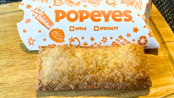 A golden-brown fried apple pie, one of Popeyes&rsquo; signature fast food desserts, sits on a wooden board in front of a Popeyes wrapper labeled &ldquo;APPLE&rdquo; and &ldquo;SPECIALTY.&rdquo;