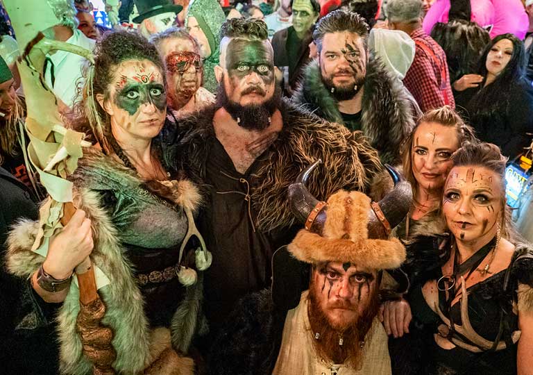 A group of people in elaborate Viking costumes and face paint pose together at a lively indoor Halloween event, surrounded by other attendees in colorful outfits.