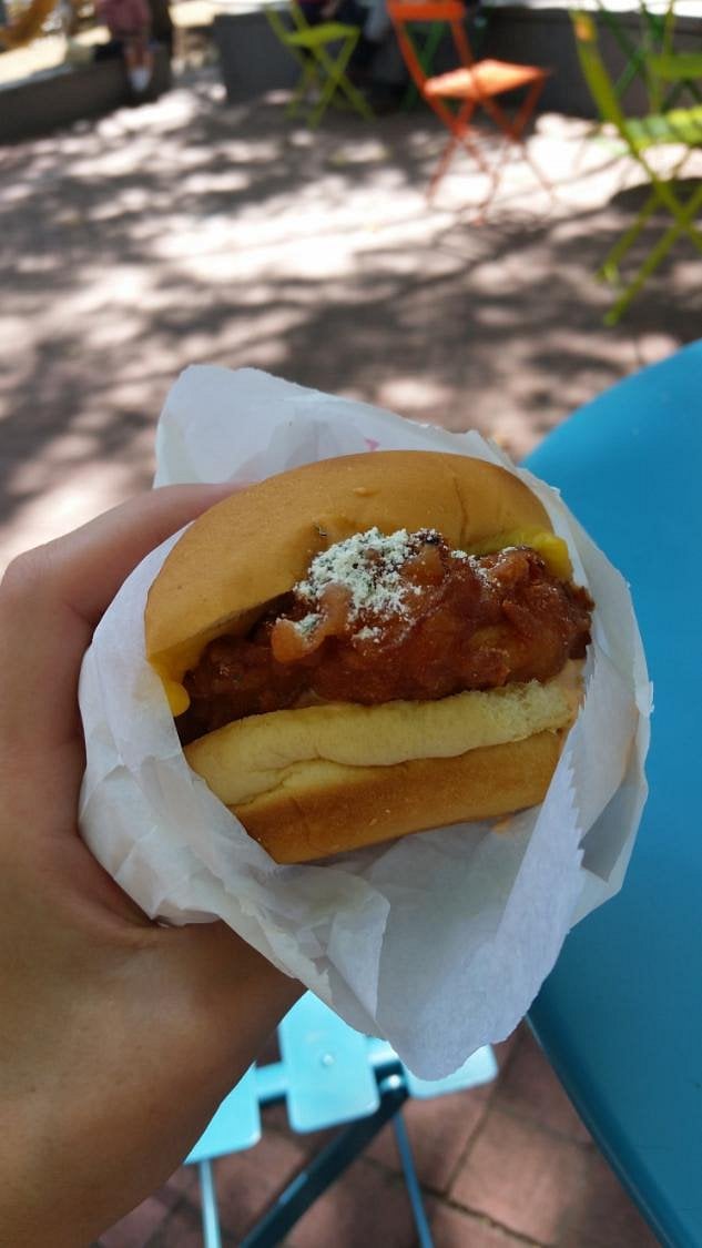 A hand holds a chili cheese hot dog in a bun, wrapped in white paper. Outdoor seating with colorful chairs and tables is visible in the background.