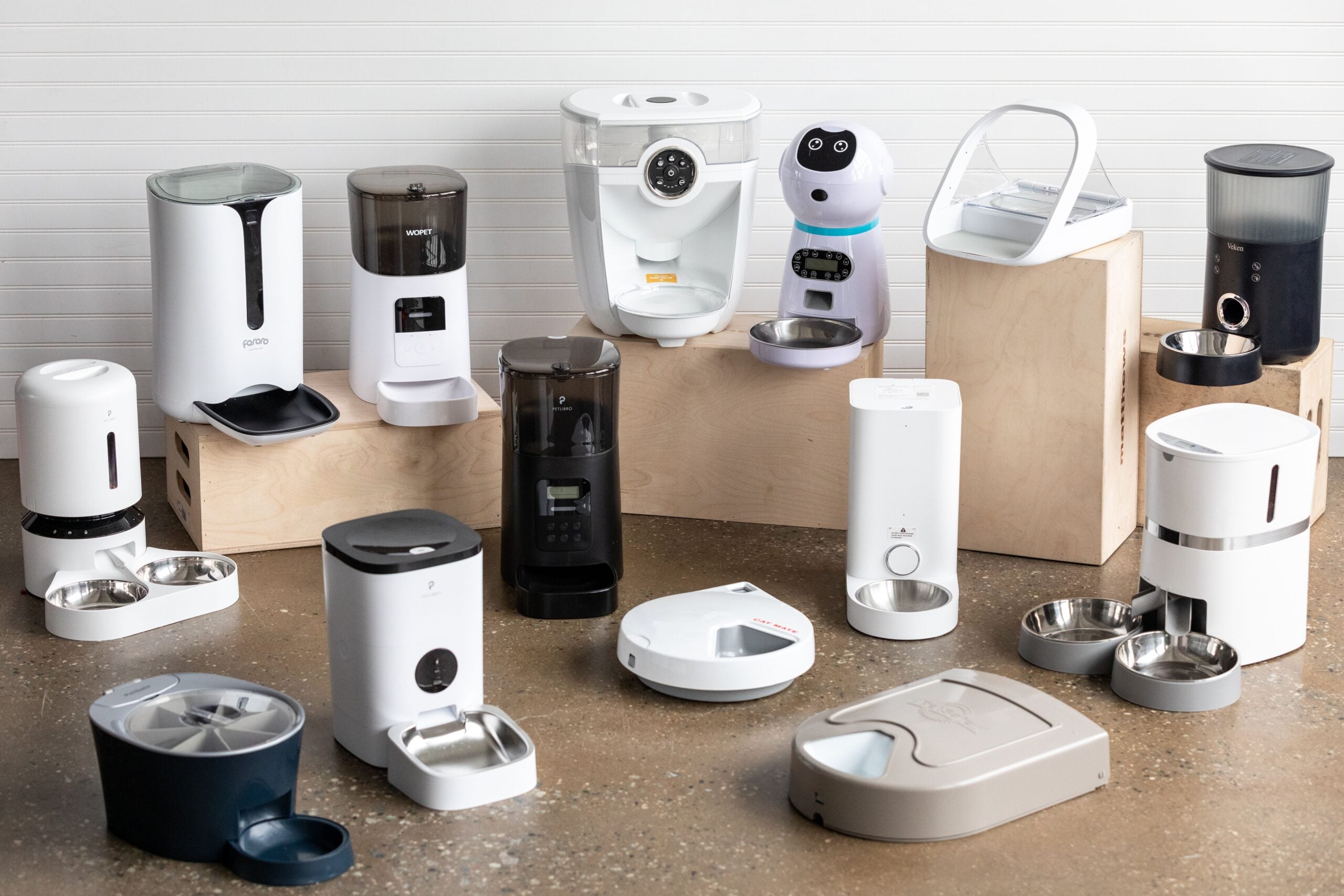 A variety of automatic pet feeders and water dispensers are arranged on wooden boxes and the floor in a bright room with a white wall.