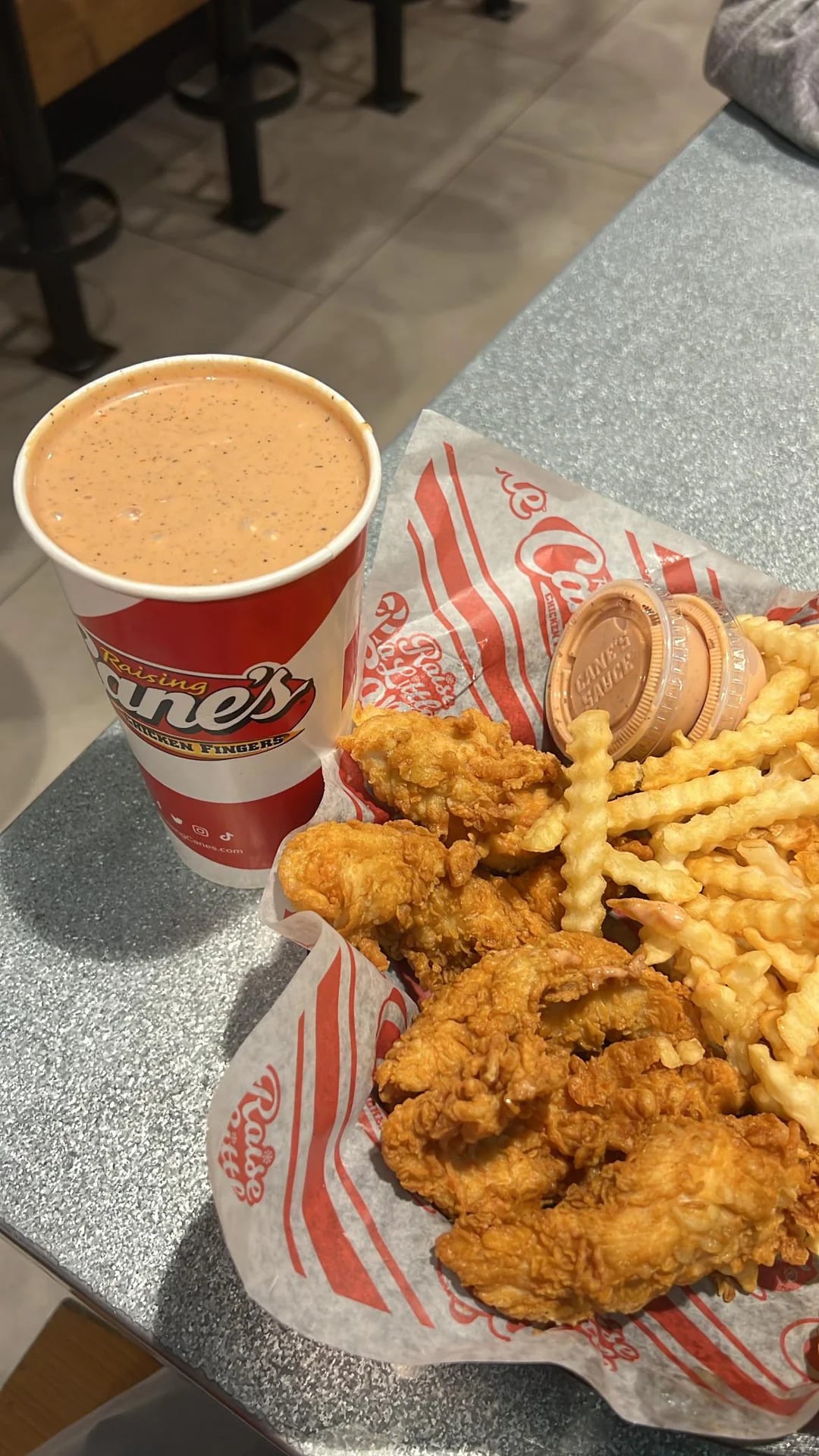 A meal with crinkle-cut fries, fried chicken tenders, two sauce cups on branded paper, and a large drink in a Raising Cane’s cup on a table—bringing the signature taste of fast-food grocery store products right to your dining experience.