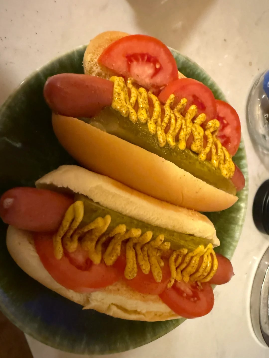 Two hot dogs in buns topped with sliced tomatoes, pickles, and zigzags of yellow mustard are served on a green plate.