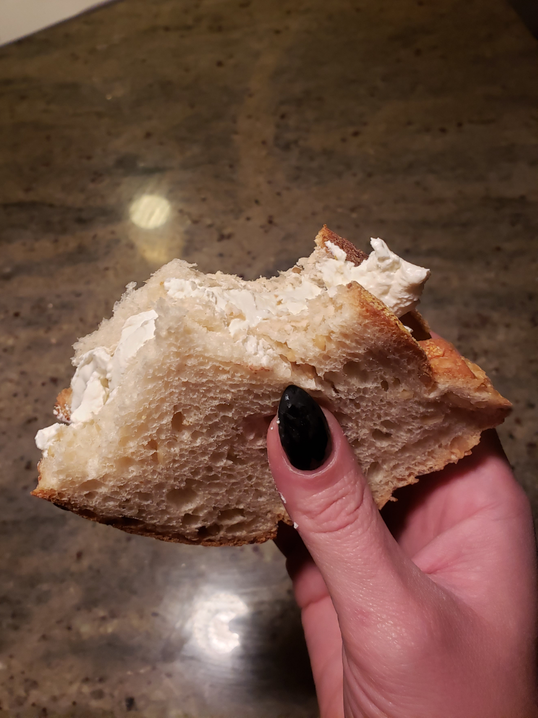 A hand with black nail polish holds a bitten piece of bread with a thick layer of cream cheese, against a brown countertop.