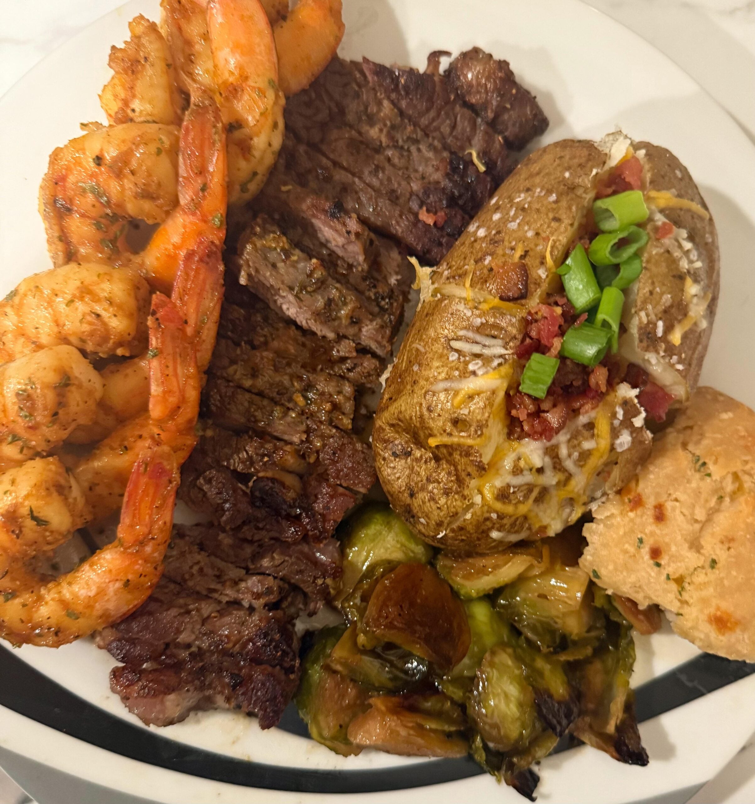 Grilled shrimp skewers, sliced steak, baked potato with cheese and green onions, roasted Brussels sprouts, and a biscuit on a white plate.