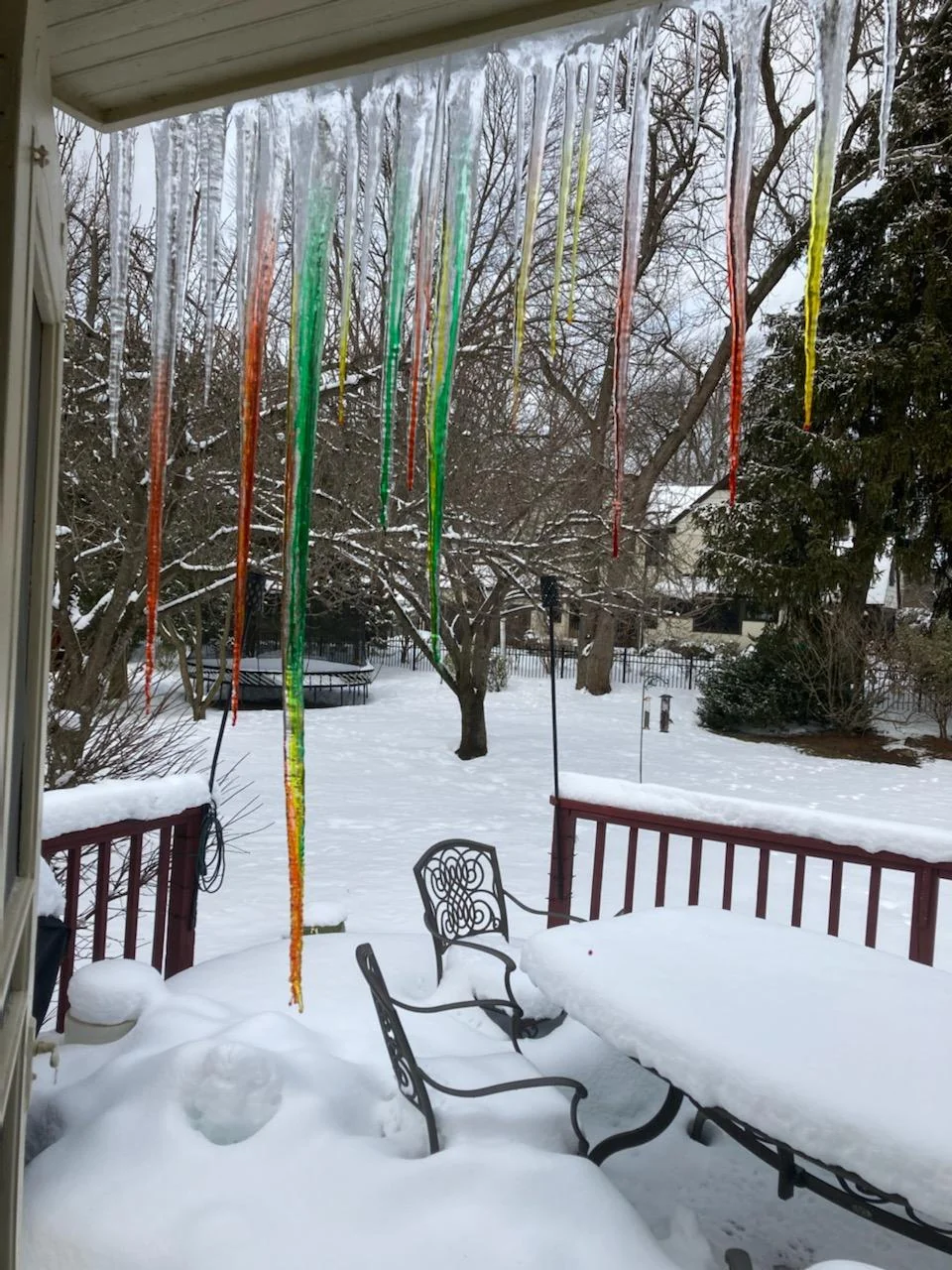Colorful icicles hang from a roof over a snowy backyard. A patio table and chairs are covered with snow, and bare trees stand in the background. The icicles are red, green, orange, and yellow.