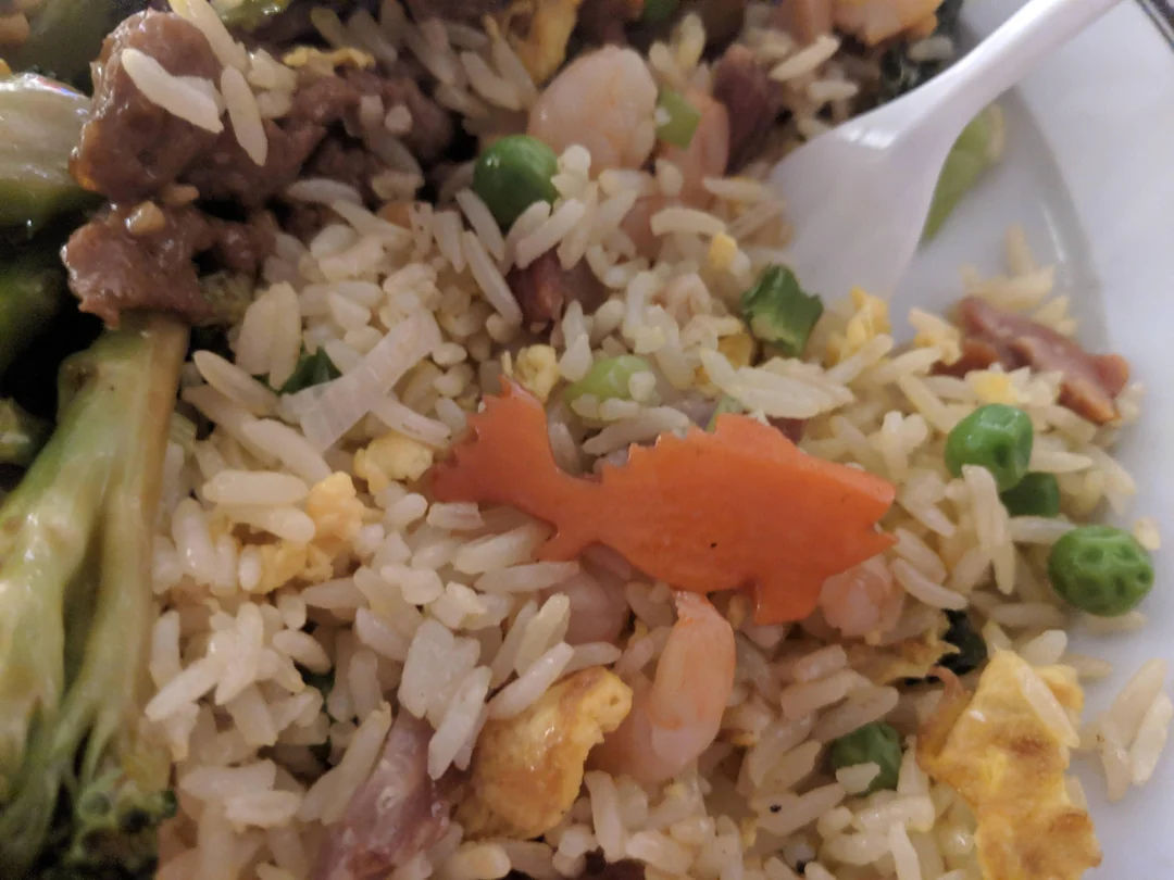 A plate of fried rice with vegetables, shrimp, eggs, and a carrot slice cut into the shape of a fish, next to a plastic fork.
