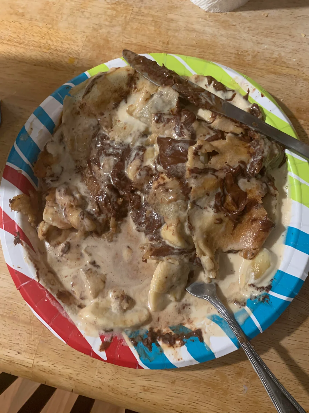 A paper plate with a serving of cinnamon roll covered in melted icing, partially eaten, with a fork and knife on the plate on a wooden table.
