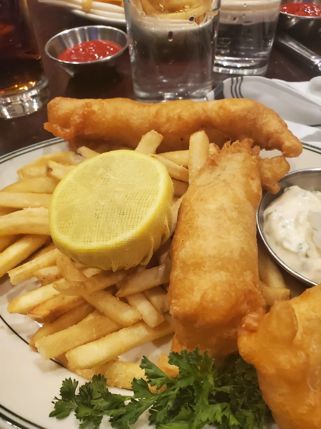 A plate of fish and chips with French fries, two pieces of fried fish, a lemon wrapped in mesh, parsley, a cup of tartar sauce, and ketchup in the background.