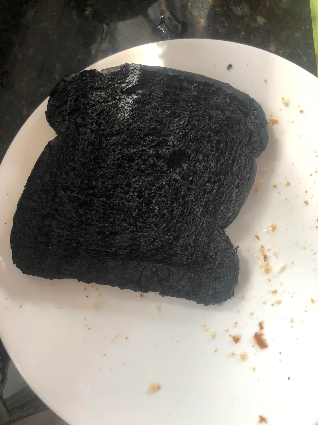 A slice of bread is burnt completely black on a white plate, with some crumbs around it.