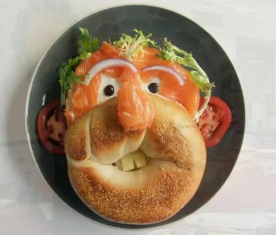 A bagel arranged on a plate to look like a face, with vegetables, smoked salmon, and cheese forming facial features such as eyes, nose, mouth, and hair.