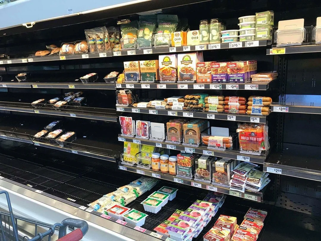 Partially empty grocery store shelves hold various packaged foods, including tofu, vegan items, and refrigerated products. Some shelves are nearly bare, while others have neatly stocked items. A shopping cart is visible in the foreground.