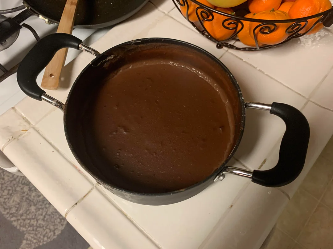 A pot filled with thick, dark chocolate pudding sits on a white kitchen counter near a fruit basket holding oranges and bananas. A wooden spatula rests in a nearby pan.