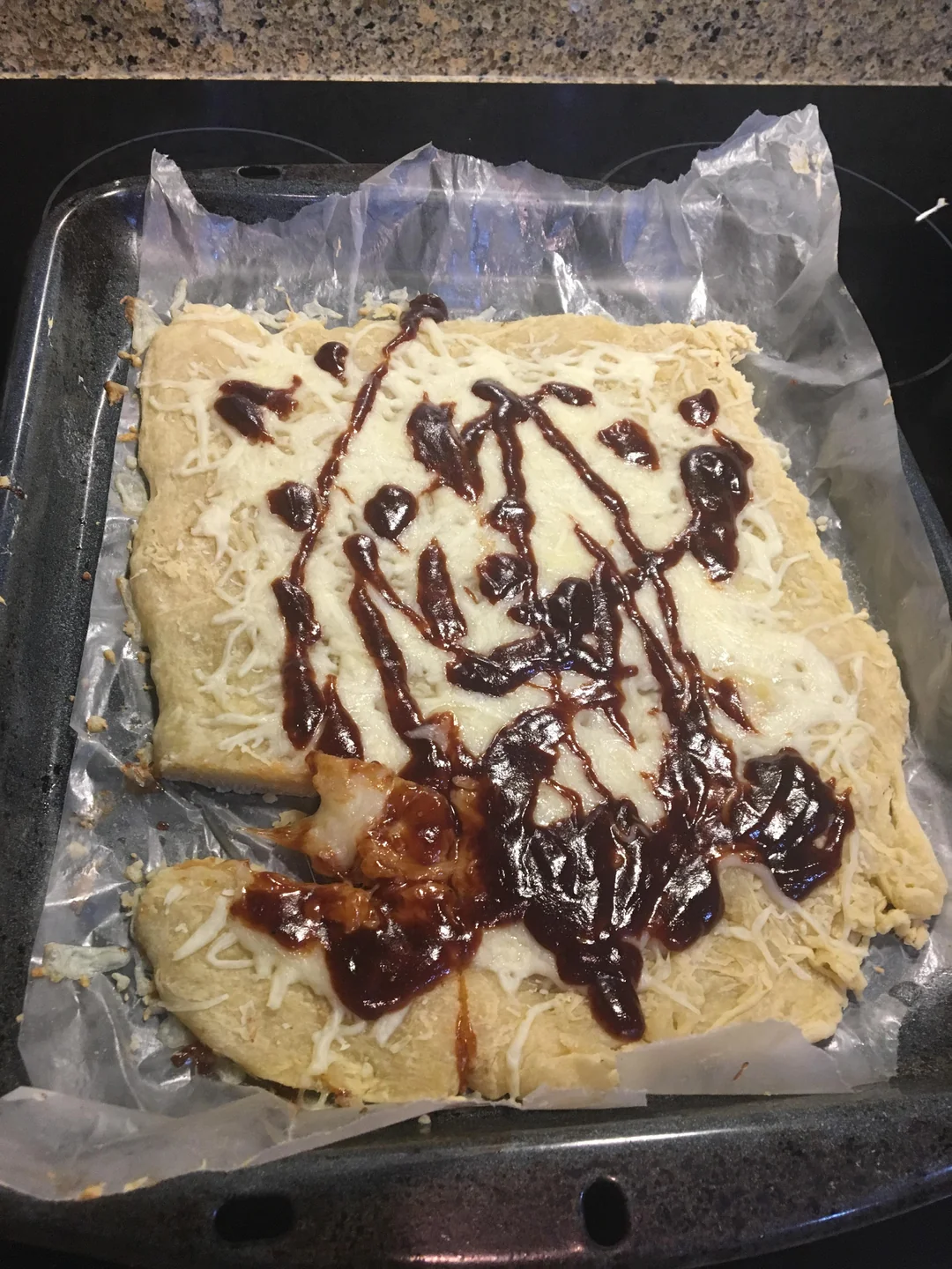 A homemade pizza with uneven edges, topped with melted cheese and streaks of barbecue sauce, sits on a parchment-lined baking tray.