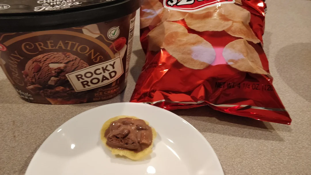 A plate with a potato chip topped with rocky road ice cream sits in front of a tub of rocky road ice cream and a bag of potato chips on a kitchen counter.
