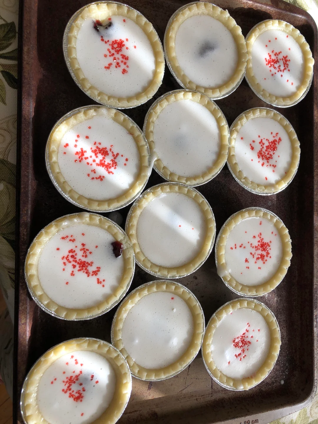 Twelve unbaked mini pies with white filling, some topped with red sprinkles, are arranged on a dark metal baking tray.