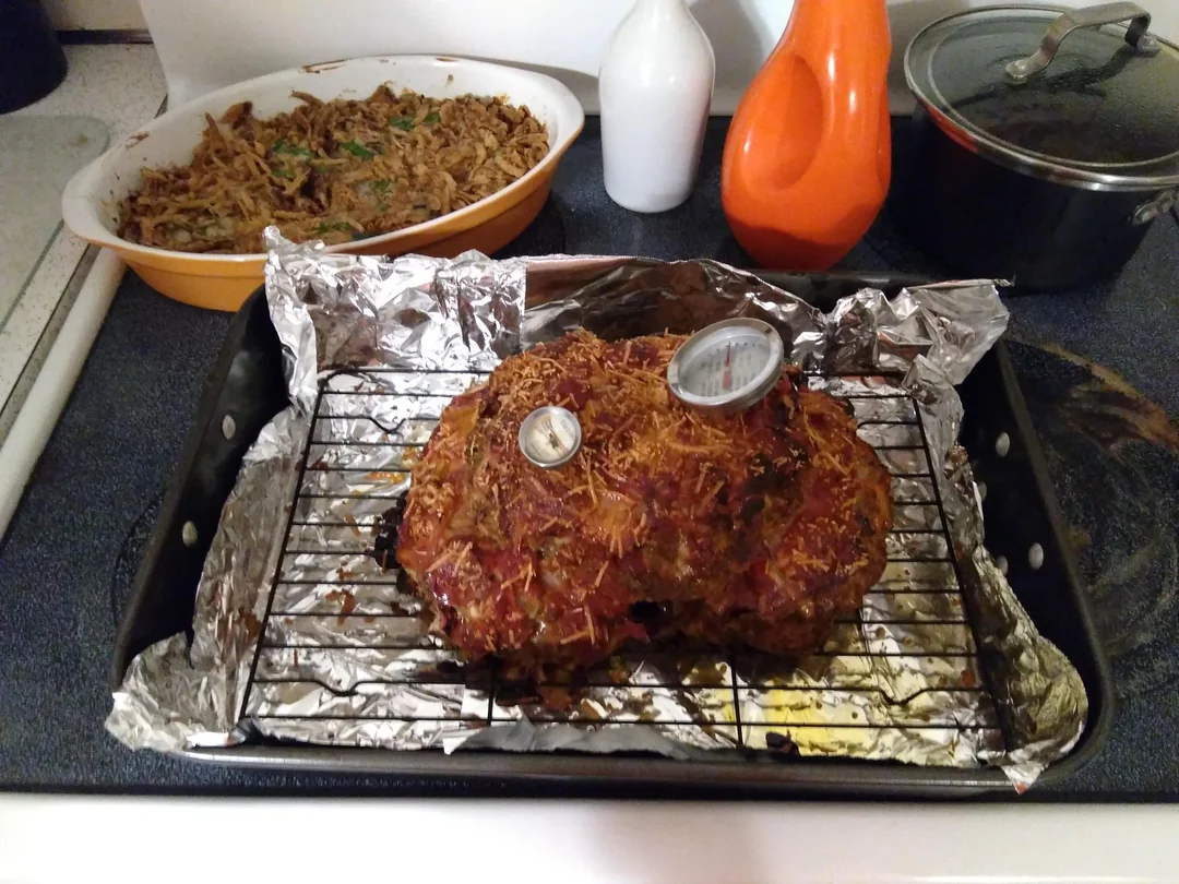 A cooked meatloaf with a thermometer sits on a foil-lined rack in a roasting pan. In the background are a dish of green bean casserole and two ceramic bottles, next to a pot with a lid on the stovetop.
