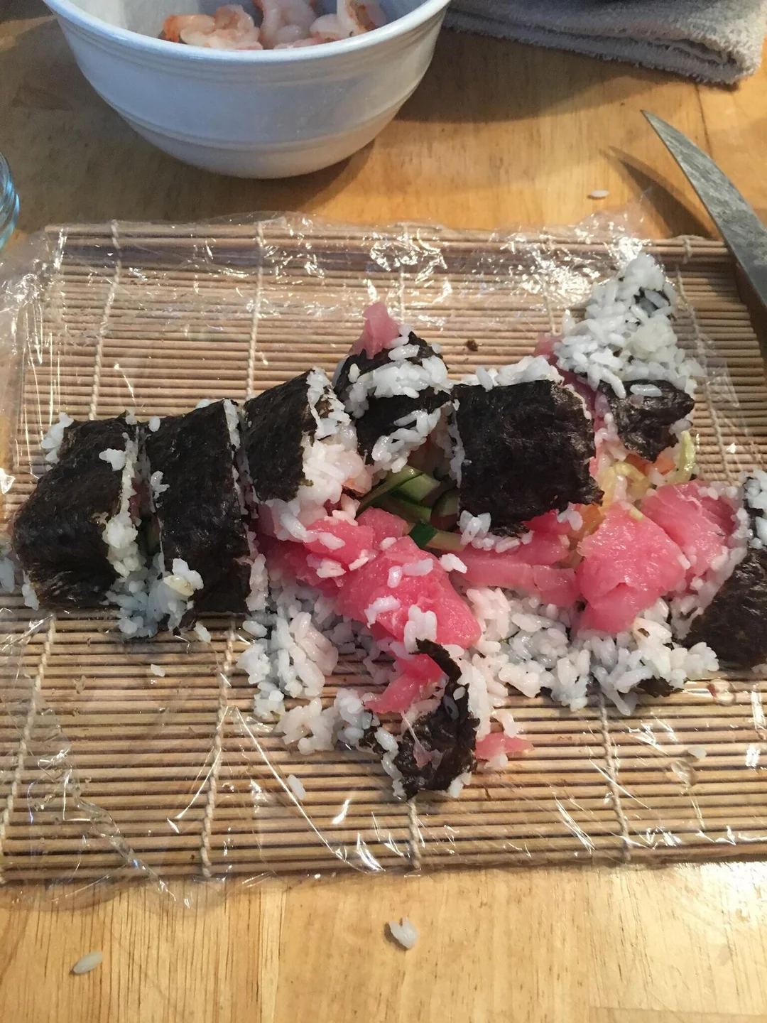 Messy homemade sushi roll with rice, seaweed, and pink fish, partially unrolled and unevenly cut, sits on a bamboo sushi mat with plastic wrap on a wooden table. A bowl of shrimp is in the background.