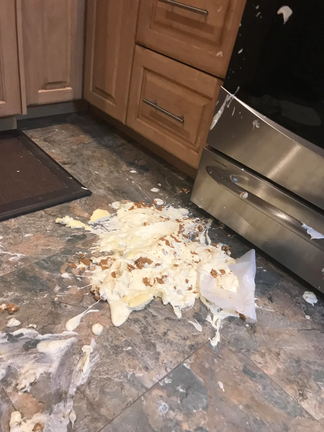 A spilled dessert with cream and crumbs is on a kitchen floor next to wooden cabinets and a stainless steel oven.