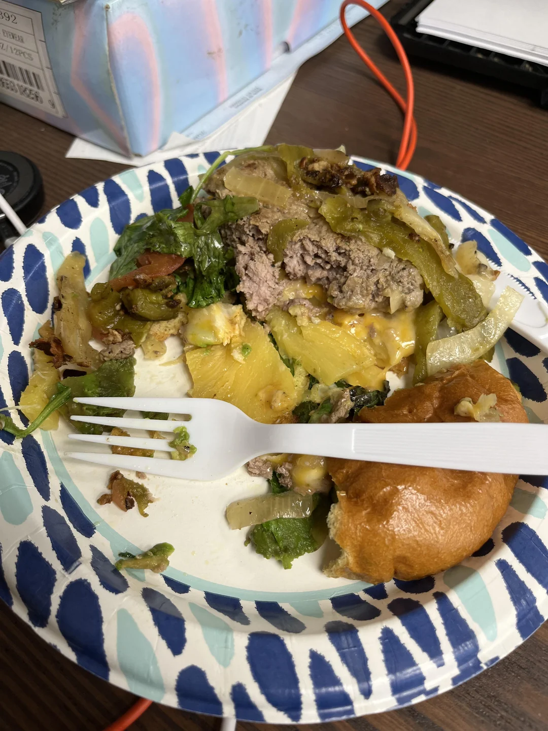 A paper plate with a half-eaten cheeseburger topped with lettuce, a plastic fork, and part of a bun, on a table with a tissue box, orange cable, and papers in the background.