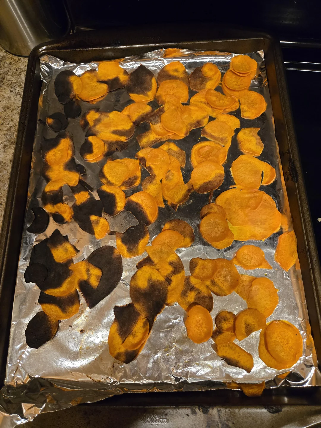 A sheet pan covered in aluminum foil holds homemade sweet potato chips, many of which are burnt black on one side, with some golden-orange chips visible. The pan is on a kitchen counter.