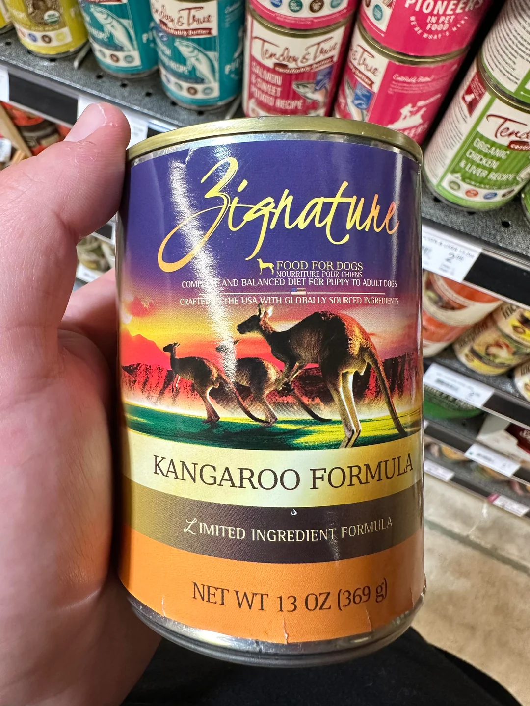 A hand holds a can of Zignature Kangaroo Formula dog food in a store aisle. The label shows kangaroos and states it is a limited ingredient formula and weighs 13 oz (369g).