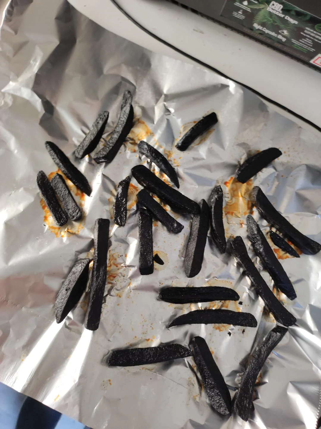 Burnt French fries scattered on a crumpled sheet of aluminum foil, with some orange-brown residue visible around them.