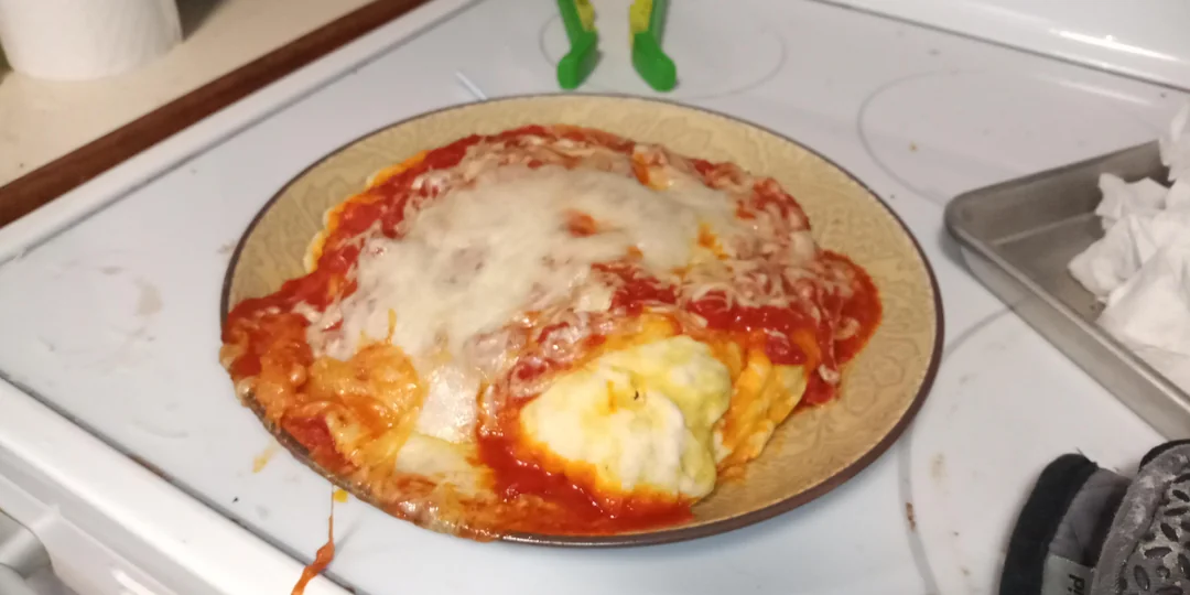 A plate of ravioli covered in tomato sauce and melted cheese sits on a stovetop next to a metal pan and some paper towels.