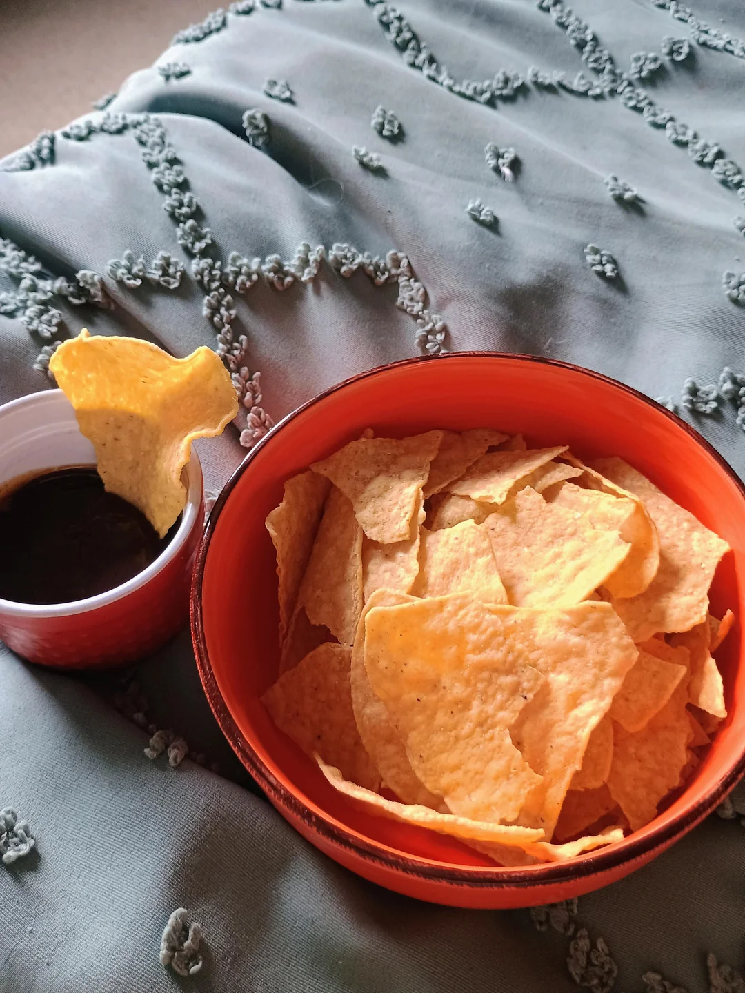 A red bowl filled with tortilla chips sits on a textured blanket next to a matching cup containing dark dipping sauce. One chip is positioned in the cup, ready to be dipped.
