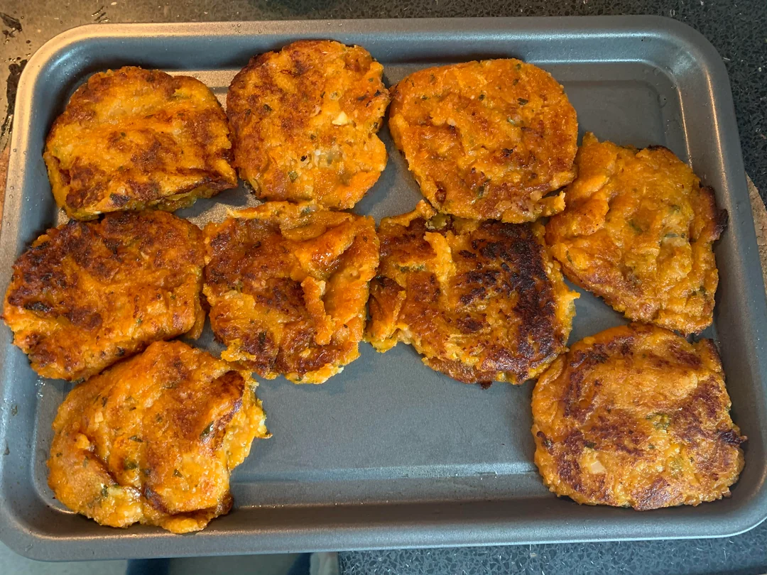 A baking tray with nine golden-brown, homemade patties or fritters, slightly uneven in shape, resting on a dark kitchen countertop.