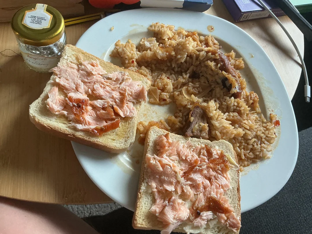 A plate with two slices of bread topped with salmon and a serving of rice with pieces of meat. A small jar and a pen are on the table nearby.