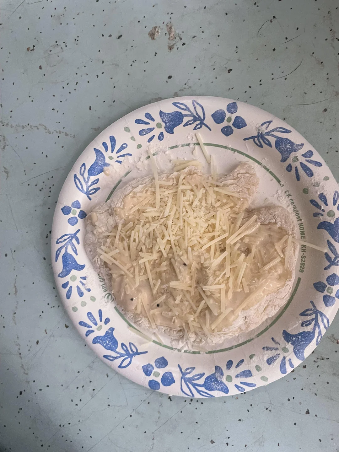 A paper plate with two pieces of white bread topped with shredded cheese and a creamy sauce, set on a worn, light blue surface with speckles.