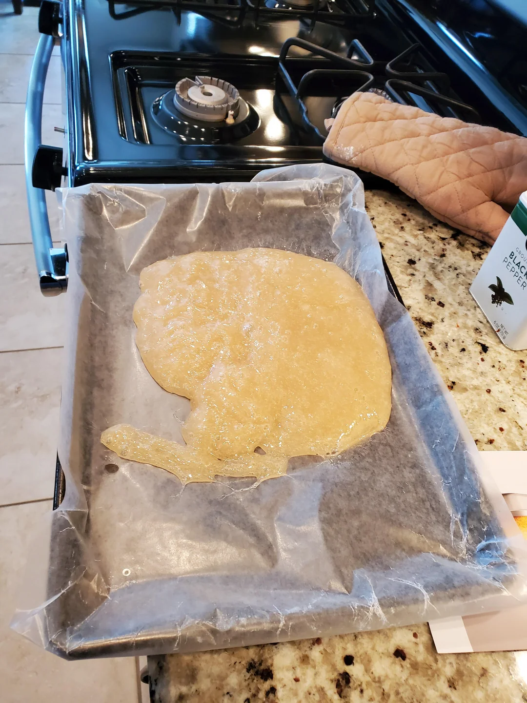A baking tray lined with wax paper holds an uneven spread of light brown batter. The tray is on a granite countertop next to a stove, oven mitt, and a green and white container.