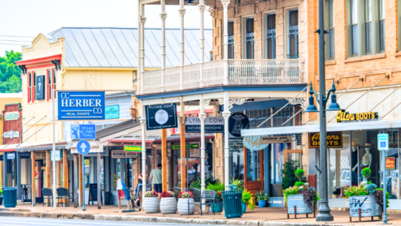 Colorful small-town street scene with old-fashioned shopfronts, various business signs, potted plants, and an ornate balcony, creating a lively and welcoming downtown atmosphere on a sunny day&mdash;perfectly capturing the charm of food towns where local eateries and specialty shops invite visitors to explore delicious flavors.