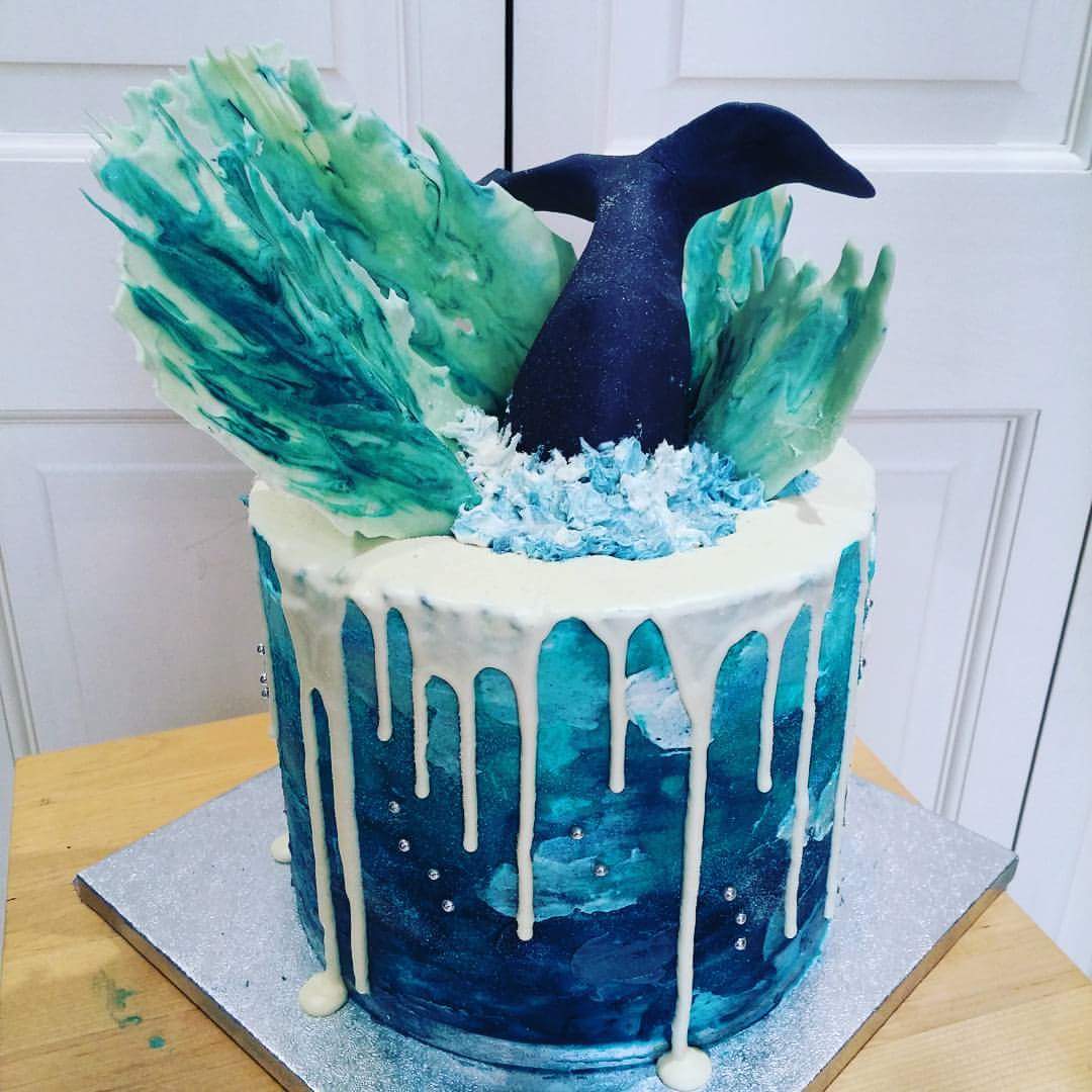 A blue ocean-themed cake with white icing drips, sugar shards resembling waves, and a black whale tail emerging from the top, set on a silver board.