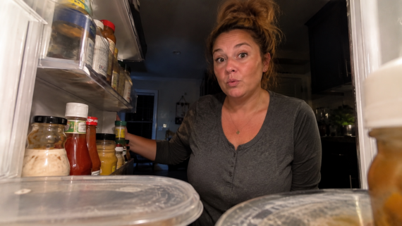 A woman with a surprised expression looks into an open refrigerator, surrounded by condiments and food containers&mdash;some of which remind her of home products people no longer buy&mdash;in a dimly lit kitchen.