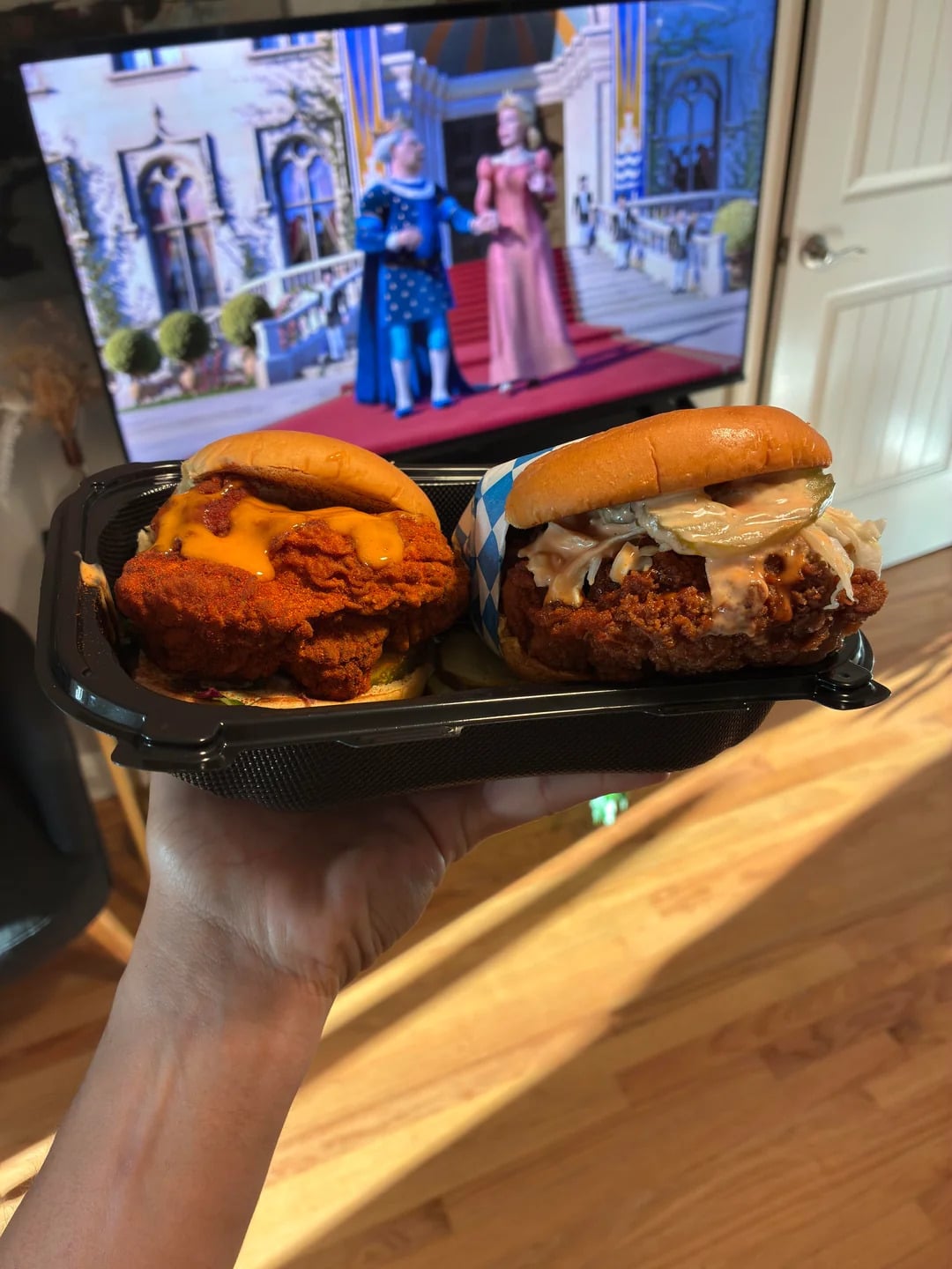 A hand holds two fried chicken sandwiches in a black tray; a TV in the background shows two animated women in front of a castle.