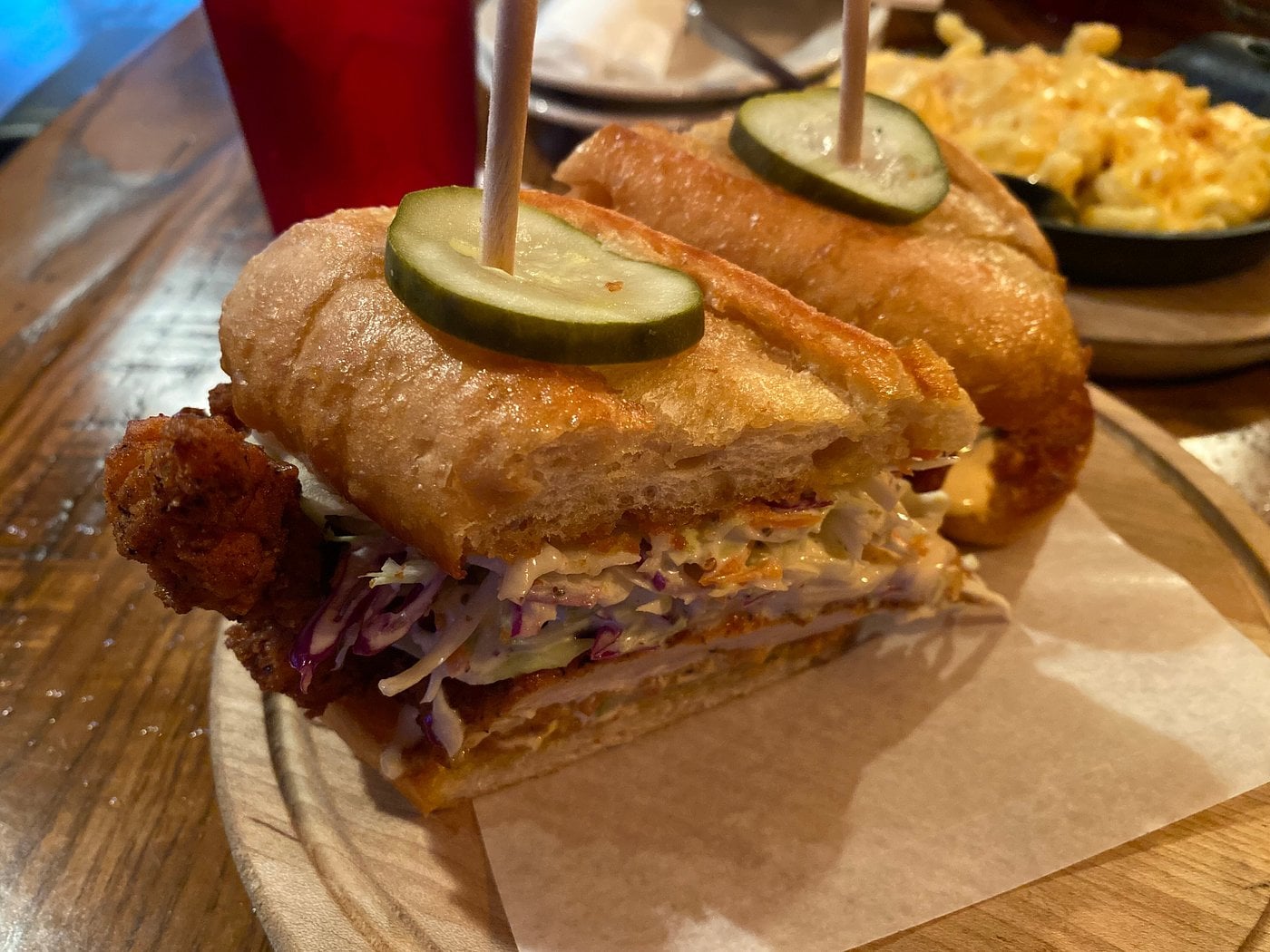 A fried chicken sandwich with coleslaw and pickle slices on a toasted bun, cut in half and served on a wooden board with a side of macaroni and cheese blurred in the background.