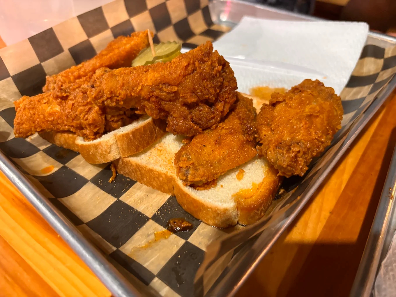 Fried chicken restaurants. Three pieces of fried chicken rest on slices of white bread with pickle chips on top, served on a metal tray lined with checkered paper and a napkin in the background.