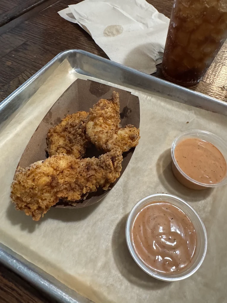 Three crispy chicken tenders on brown paper, served with two cups of dipping sauce on a metal tray lined with parchment, with a drink and napkin in the background.