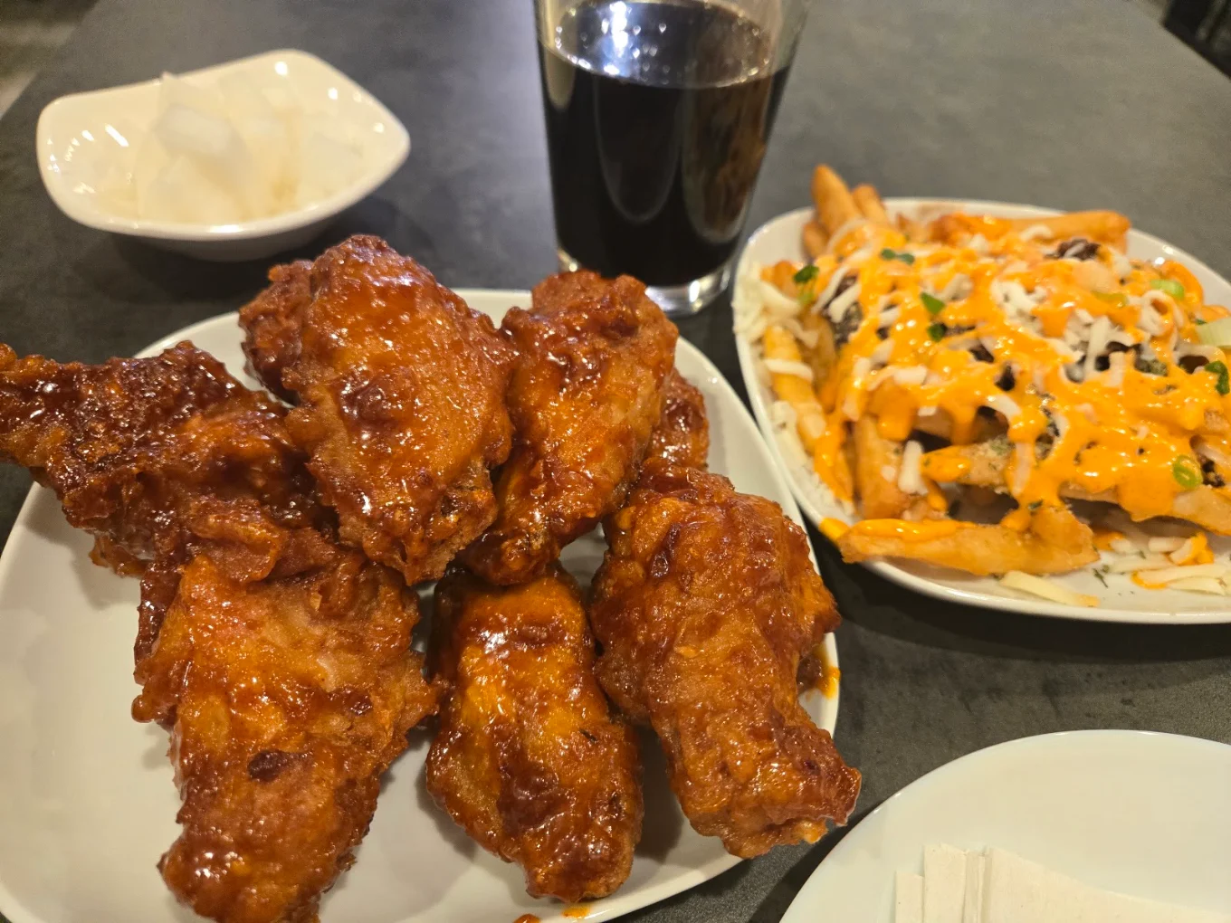 A plate of glazed fried chicken wings, loaded fries with cheese and sauce, a glass of dark soda, and a bowl of pickled radish sit on a dark table.