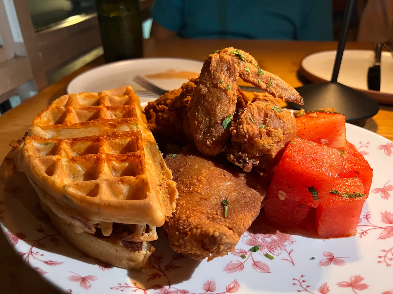 A plate of golden waffles, crispy fried chicken, and cubes of fresh watermelon is served on a floral-patterned plate on a wooden table under warm lighting.