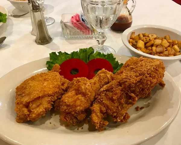 A plate with three pieces of fried chicken, two slices of red tomato, and lettuce. There is a glass of water, a bowl of diced potatoes, and condiments in the background on a white tablecloth.