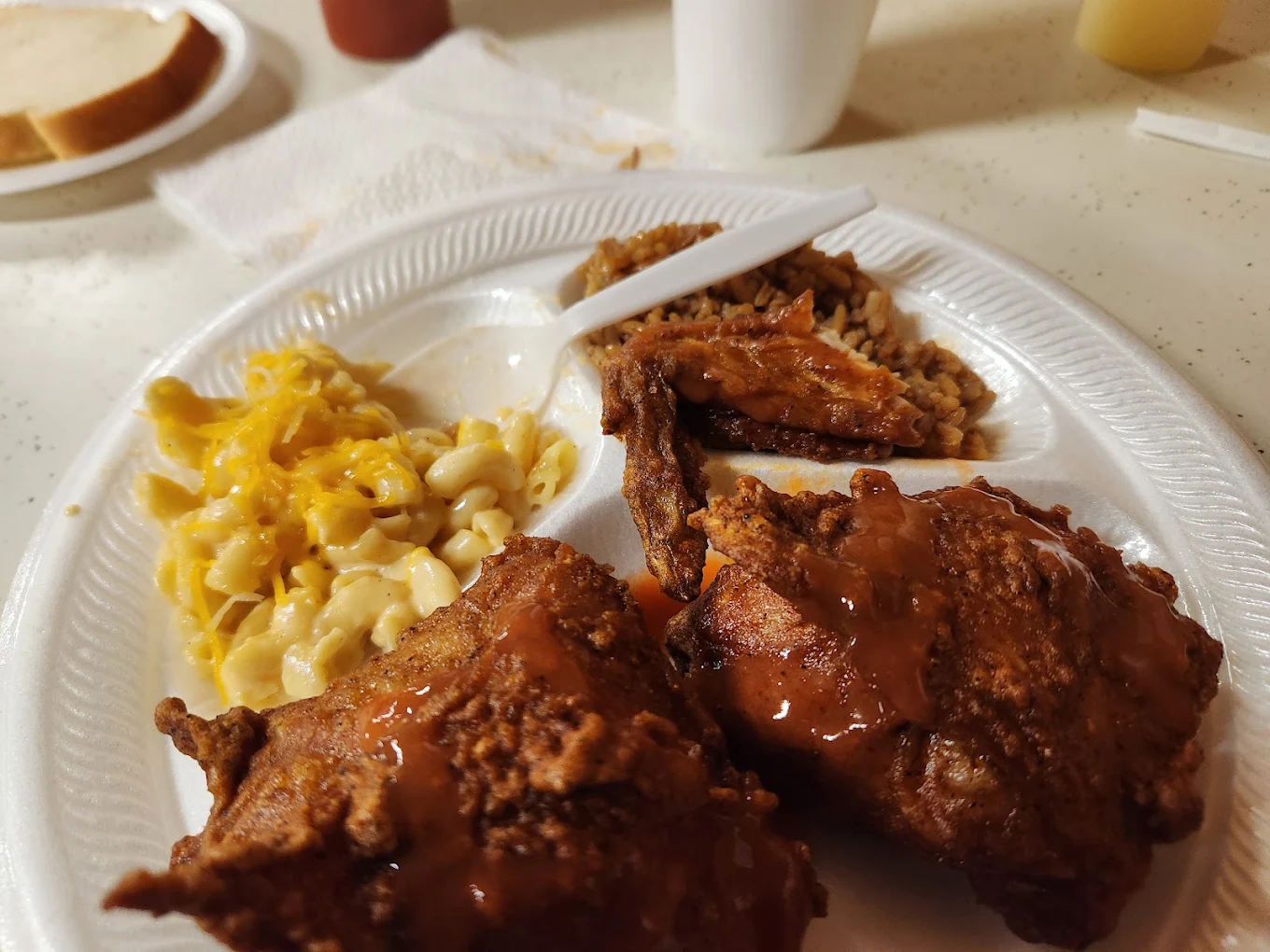 A styrofoam plate with macaroni and cheese, two pieces of saucy fried chicken, and a serving of rice with gravy. A plastic fork, cup, and a slice of bread are visible in the background.