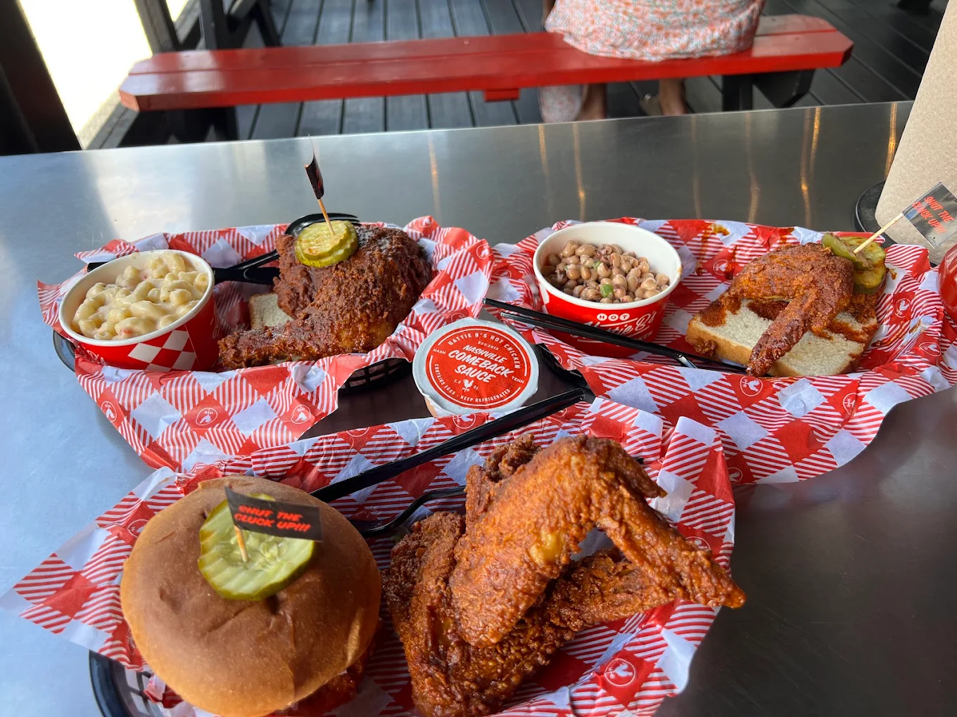 Three trays of Nashville hot chicken meals with pickles, mac and cheese, and beans sit on a metal table with red checkered paper, seen in an outdoor dining area. A person sits at a picnic table in the background.