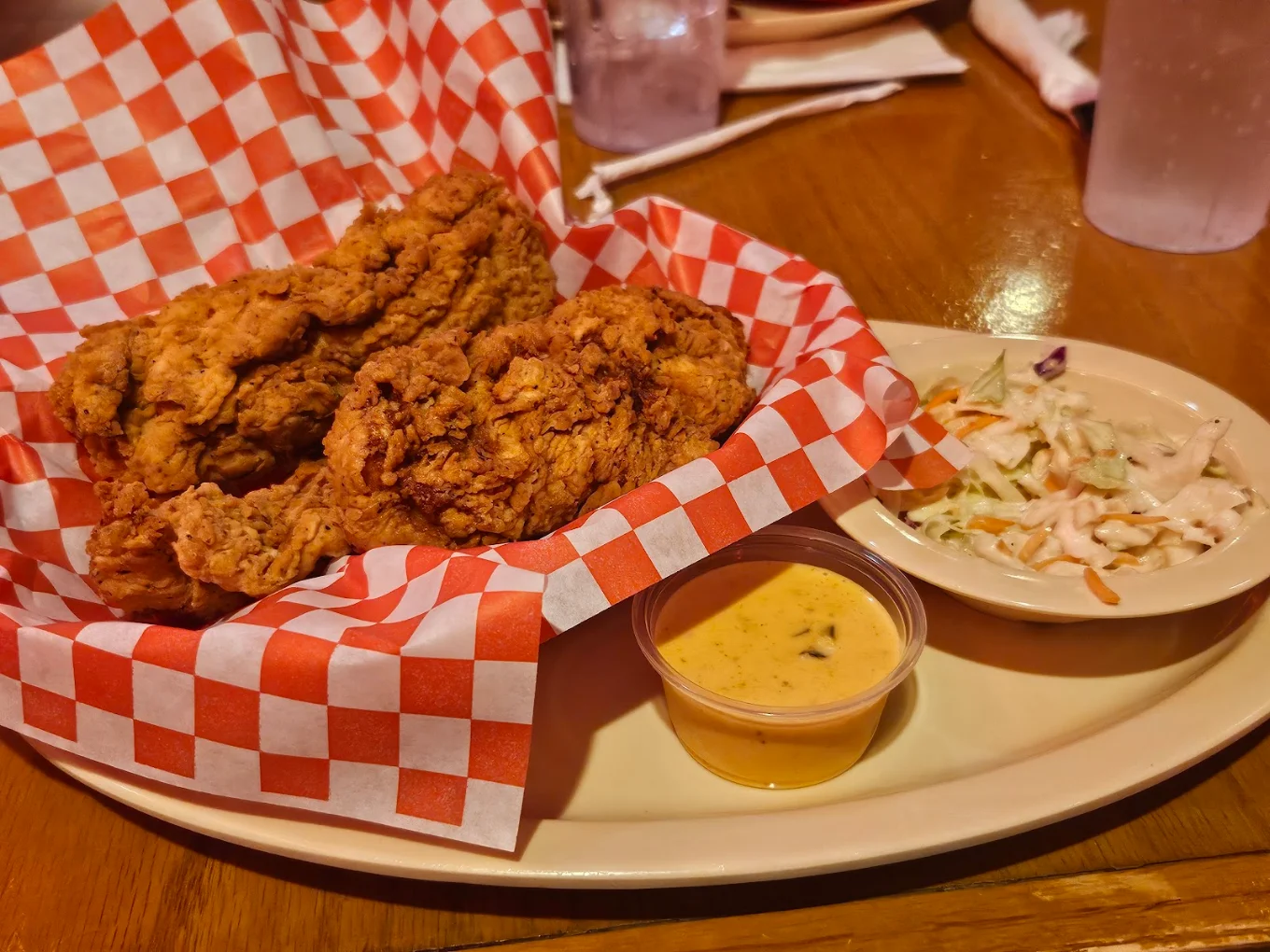 Basket of fried chicken on red checkered paper, served with coleslaw and a small cup of dipping sauce on a beige plate, set on a wooden table.