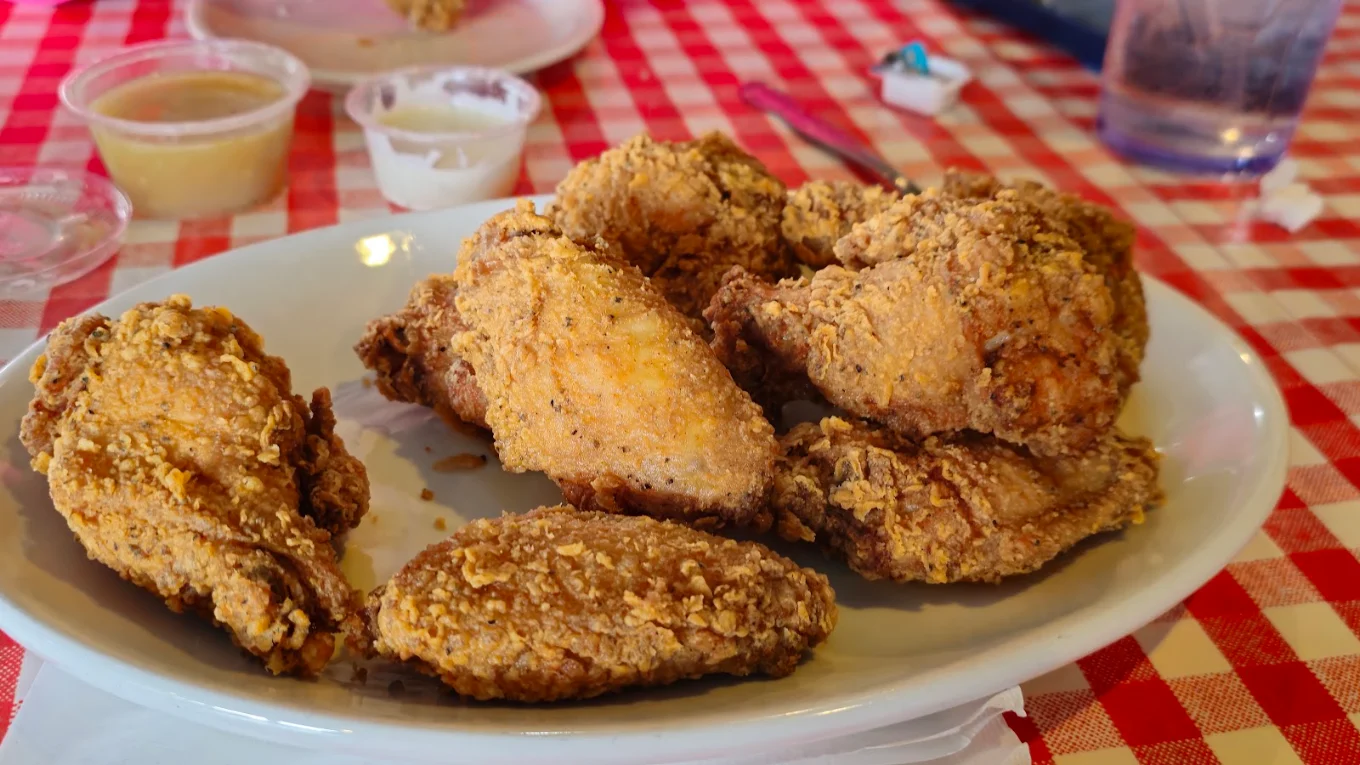 A plate of crispy fried chicken pieces sits on a red and white checkered tablecloth, with dipping sauces and a partially eaten plate in the background.
