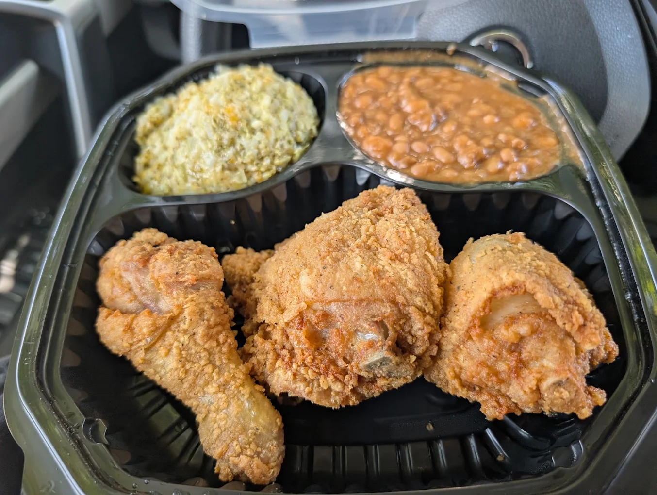 A takeout container with three pieces of fried chicken, baked beans, and a serving of stuffing or dressing, all in separate compartments.